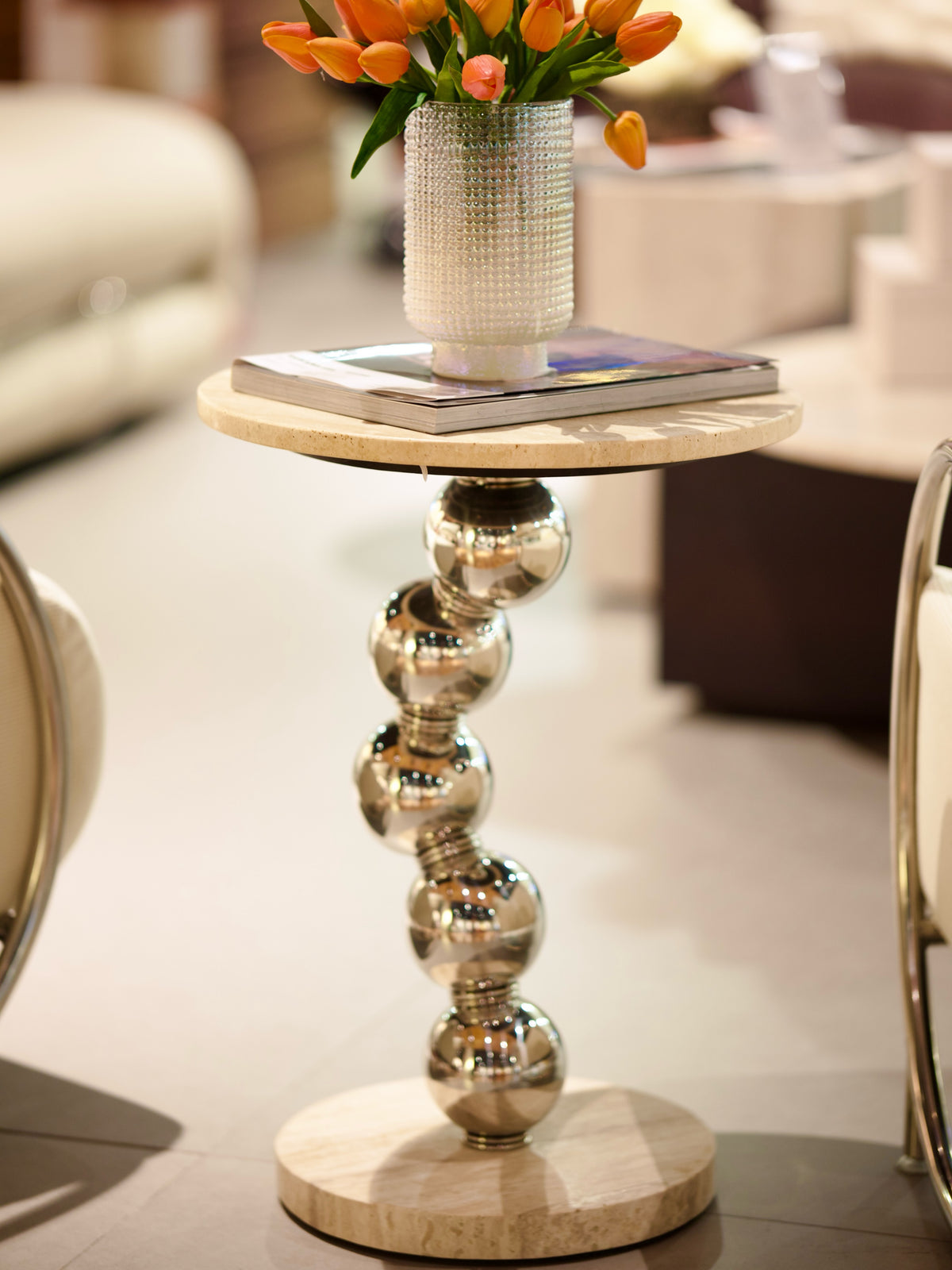 A round side table with a polished silver ball-column pedestal and a light beige stone top, styled with a textured white vase of orange tulips and a magazine in a modern interior setting.